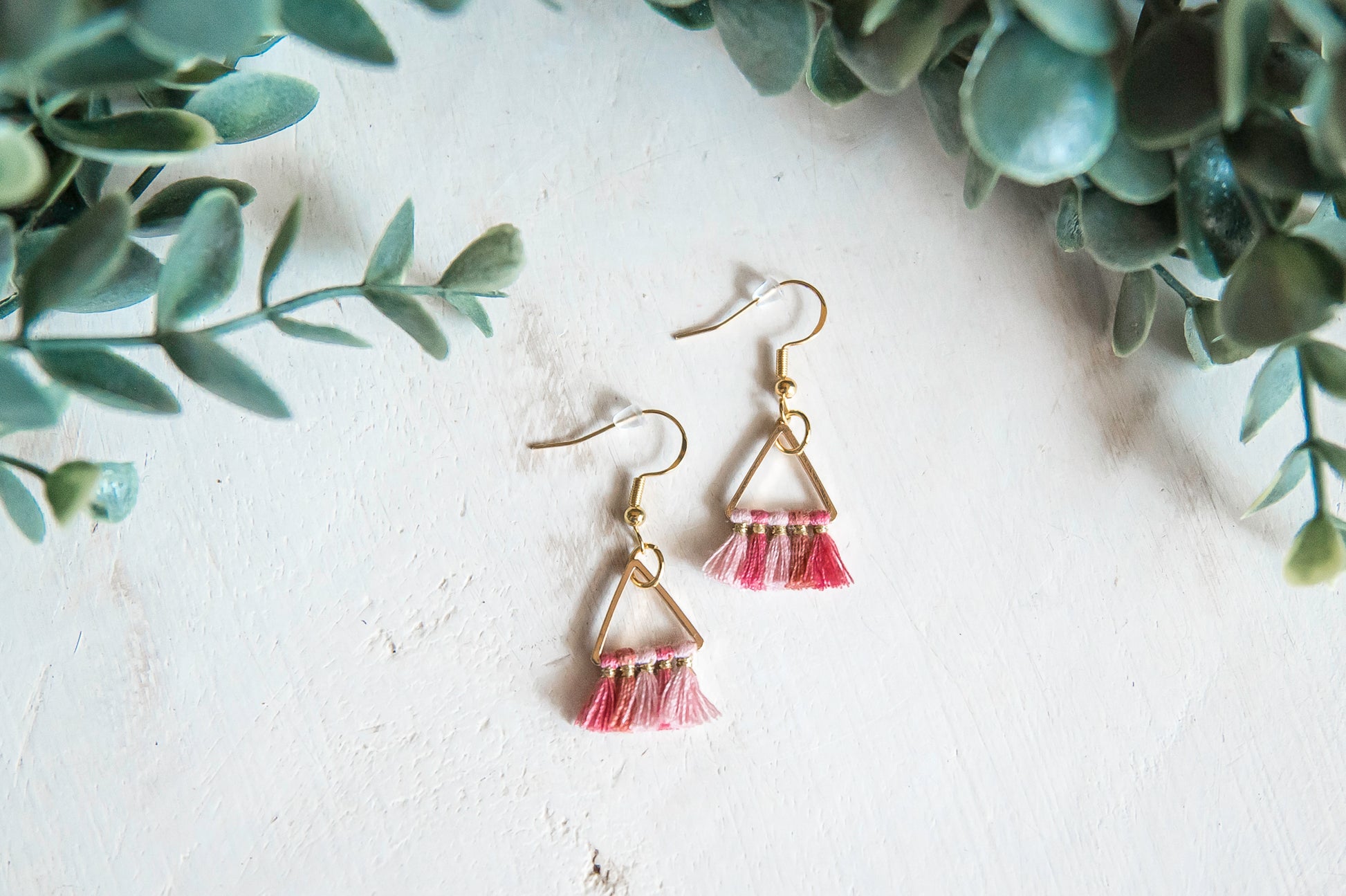 Petite gold triangle earrings with gold hypoallergenic hooks and rubber back pieces. Shades of pink thread dangle from triangular piece. Set against white background with greenery.