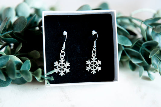 pair of silver snowflake earrings in a black velvet box against some green eucalyptus leaves