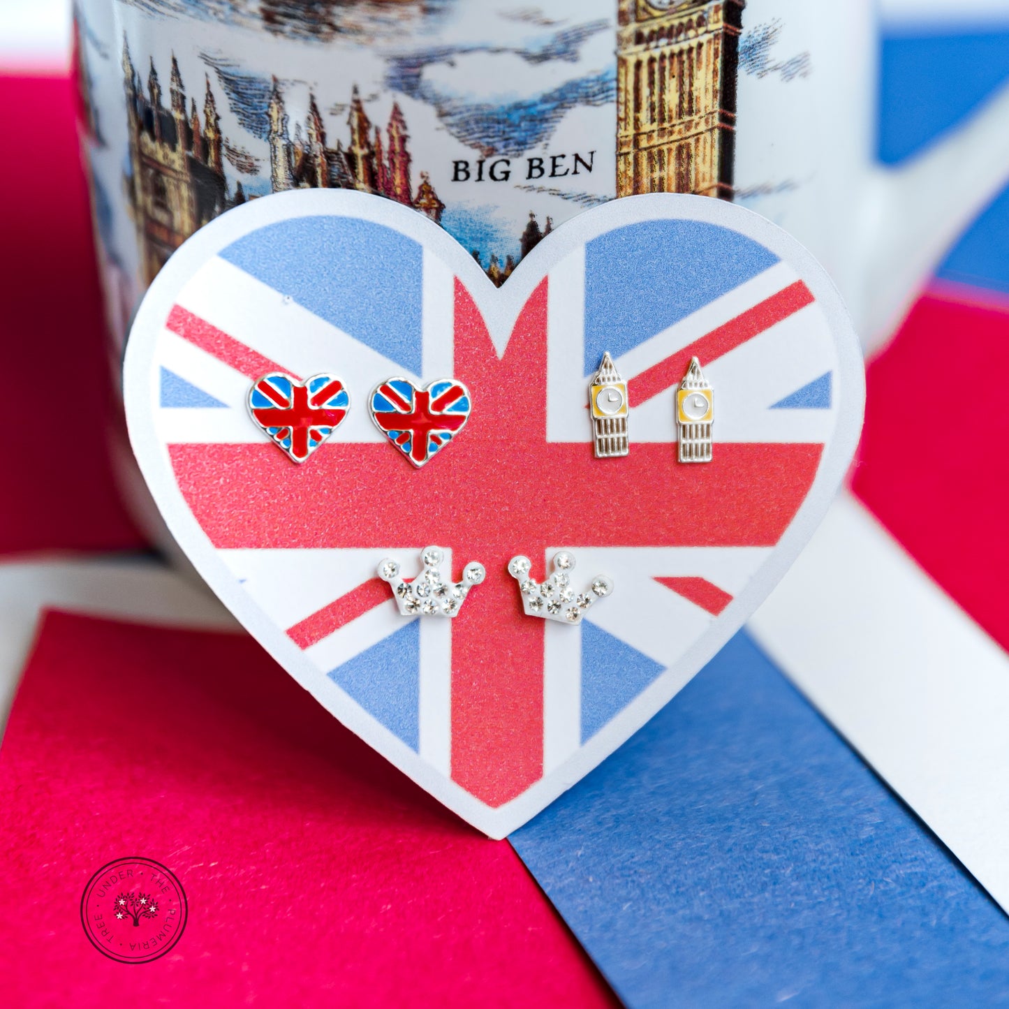 Flag of Great Britain is in a heart shape and printed with red, white, and blue. It is an earring holder for 3 pairs of earrings - one as heart Great Britain earrings, one as clock towers that are reflective of Big Ben, and one of sparkly crowns. Set against an image of the Houses of Parliament and on red, white, and blue tablecloth