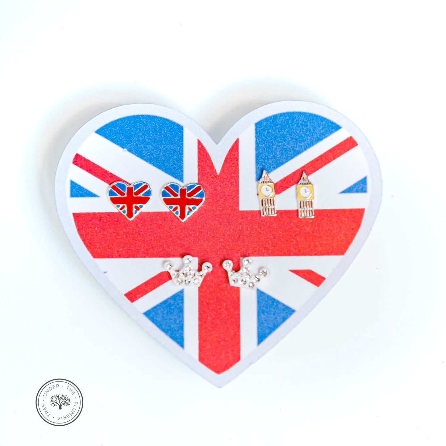 White background with an earring paper holder in the shape of heart with 3 pairs of earrings - Union Jack love with a flag of Great Britain in the shape of a heart, mini Big Ben clock towers, and sparkly silver crown earrings