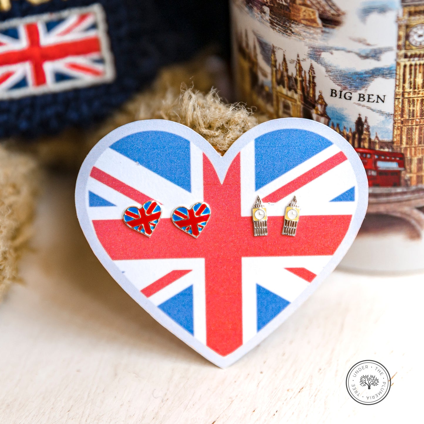 2 sets of earrings on a heart cutout of the Union Jack and set against a stuffed bear representing Great Britain and London; pairs of earrings include the Union Jack in a heart shape and Big Ben clock towers - perfect for travel, London, and everything UK