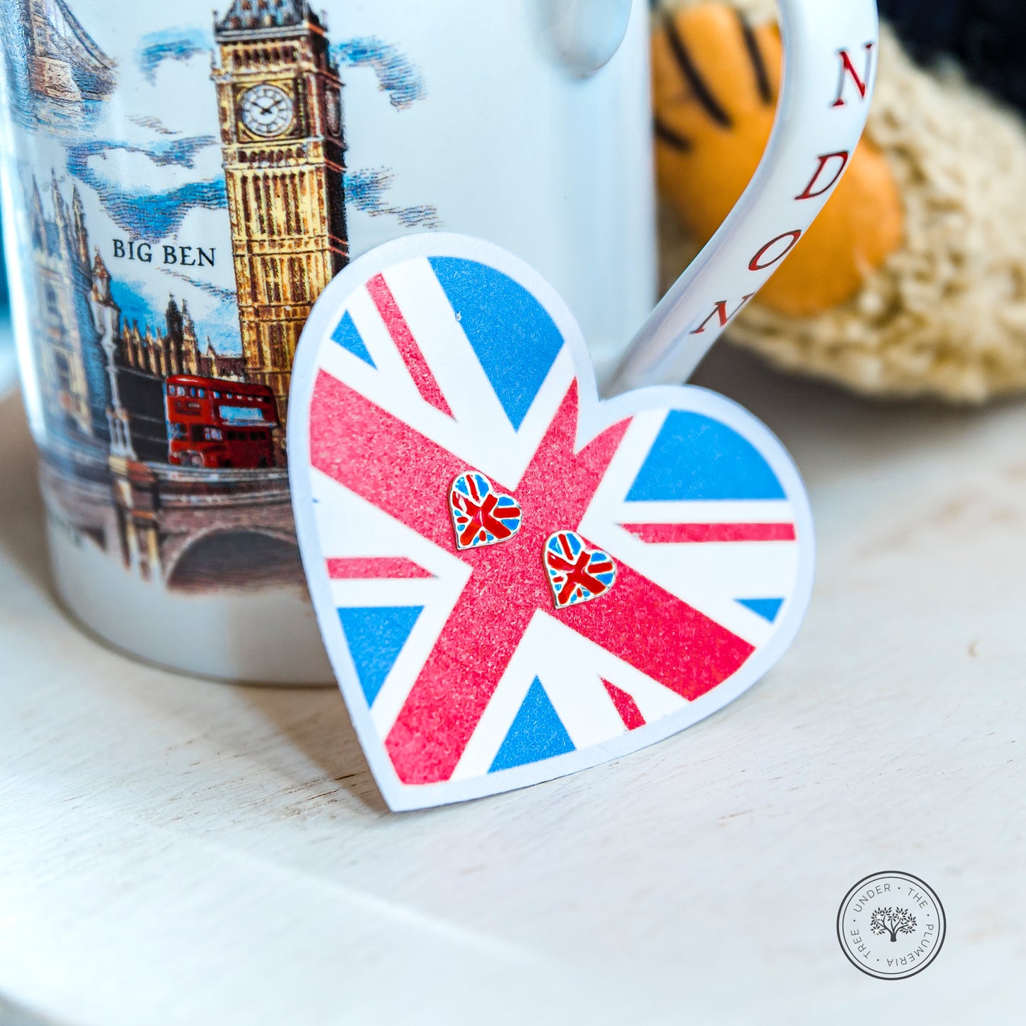 Pair of earrings in the shape of  heart and with the flag of Great Britain, set against a heart of Great Britain and resting on a London mug showing Big Ben and a London city red double decker bus